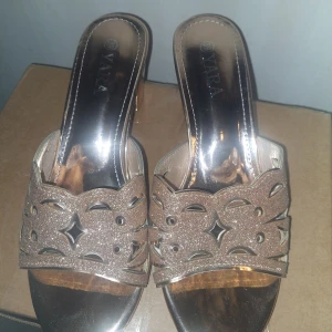 Glittriga roseguldiga skor från Yara - strl 39 - Använda en gång! I mycket bra skick.Snygga sandaler i guld med glittrig ovandel och utskurna detaljer. Skorna har öppen tå, blockklack och en elegant finish som verkligen sticker ut. Perfekta för dig som vill ha något extra till outfiten. Klackhöjd är låg och sulan är platt framtill.