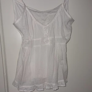Vit randig baby doll top - Söt vit blus med tunna vertikala ränder, knappar framtill och volangkant nertill. Blusen har v-ringning och är ärmlös med spetsdetalj längs halsringningen. Perfekt för varma dagar och en lätt, luftig känsla.