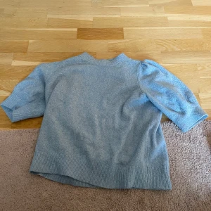 Ljusblå stickad tröja med puffärm - Superfin ljusblå stickad tröja med korta puffärmar och ribbade muddar. Perfekt för dig som vill ha en enkel men ändå trendig look. Tröjan har en rund halsringning och är mjuk och skön att bära.