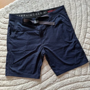 Mörkblå shorts Wrangler Terrain Gear - Snygga mörkblå shorts från Wrangler Terrain Gear med midwaist passform. De har praktiska fickor fram och bak samt ett justerbart bälte med snörning i midjan. Perfekta för sommardagar och outdoor-aktiviteter. Materialet känns lätt och smidigt.