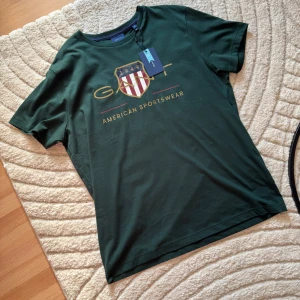 Mörkgrön GANT t-shirt med tryck XL - Snygg mörkgrön t-shirt från GANT i bomull med klassiskt tryck framtill och sköldlogga i rött, vitt och guld. Modellen är kortärmad med rund halsringning och relaxed fit. Perfekt för dig som gillar stilren och sportig vibe.