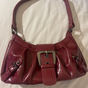 Y2k väska - Snygg vinröd Y2k axelväska i glansigt skinnliknande material med bredt spänne framtill och coola bältesdetaljer på sidorna. Väskan har dragkedja upptill och ett rymligt fack. Perfekt för dig som gillar statement-accessoarer.