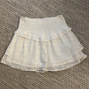 Vit volangkjol från Gina Tricot - Supersöt vit kortkjol från Gina Tricot med bred smock i midjan och dubbla volanger. Kjolen har ett diskret geometriskt mönster i tyget och är perfekt för en fräsch och somrig look. Lätt och luftig, passar perfekt till sneakers eller sandaler.