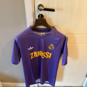 Retro Lila Real Madrid Adidas fotbollströja - Retroinspirerad lila Real Madrid fotbollströja från Adidas med klassiska vita ränder på axlarna, gul ZANUSSI-tryck på bröstet och klubbmärke broderat. Tillverkad i lätt och ventilerande polyester, perfekt för match eller träning. Kortärmad med V-ringad krage.