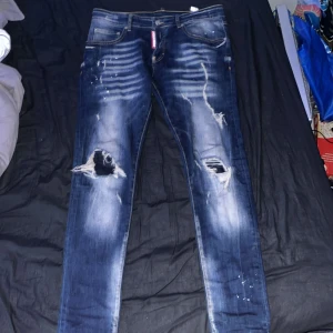 Dsquared2 blå slim fit jeans herr - Säljer ett par blå skinny jeans från Dsquared2 med slitningar, hål på knäna och coola färgstänk. Jeansen har klassisk femficksdesign, låg midja och Dsquared2-logga bak. Tillverkade i bomull med stretch för skön passform.
