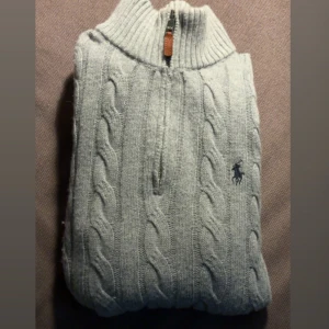 Grå kabelstickad tröja Polo Ralph Lauren - Kabelstickad grå tröja från Polo Ralph Lauren med halv dragkedja och klassisk broderad logga på bröstet. Tröjan har ribbade muddar, hög krage och är långärmad. Perfekt för dig som gillar stilren och tidlös design.