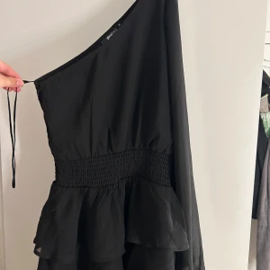 Svart oneshoulder volangklänning - Supersnygg svart kortklänning från Gina Tricot med oneshoulder och volangdetaljer. Klänningen har smockad midja och ett lager av tunna volanger nedtill.  Endast använd på 1 gala 1gång ❤️🙏🏻