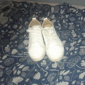 Vita sneakers med nitar och röd sula - Snygga vita sneakers med coola nitar på tån och klassisk snörning. Skorna har en unik röd sula som sticker ut och ger extra edge. Tillverkade i skinnliknande material och har en rund tå. Perfekta för dig som vill ha något annorlunda i din sneaker-samling. Obs 2 taggar saknas på skorna därav de låga priset