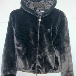Svart pälsjacka med huva från Fila - Mjuk och fluffig svart pälsjacka från Fila med stor logga på ryggen och dragkedja framtill. Jackan har huva, sidofickor och en clean look som passar perfekt till streetwear. Snygg och varm för kyliga dagar.