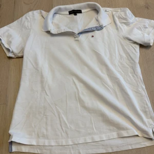 Vit piké från Tommy Hilfiger - Klassisk vit piké från Tommy Hilfiger med liten broderad logga på bröstet. Kragen har en snygg detalj med blåvita ränder på insidan. Perfekt för en clean och stilren look.