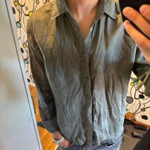 Olivgrön linneskjorta från Sandö - Snygg olivgrön skjorta från Sandö i 100% linne. Skjortan har klassisk krage, knäppning framtill och långa ärmar. Perfekt för en avslappnad men stilren look. Passar lika bra till jeans som till shorts.