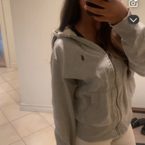 Grå sipp hoodie från Ralph Lauren - Säljer en grå hoodie från Ralph Lauren med dragkedja och luva. Tröjan har den klassiska loggan broderad på bröstet, fickor framtill och är i mjuk bomull. Perfekt för chill dagar och enkel att matcha med allt. [OBSSSS armarna är lite slitna kan visa tydligare bilder privat!] 