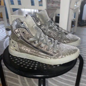 Silvriga sneakers med ormmönster - Unika silvriga sneakers med orm mönster och coola detaljer som dragkedja på sidan och snörning framtill. Skorna har en högre modell och platt sula, vilket ger en edgy look. Perfekta för dig som vill sticka ut med din stil.