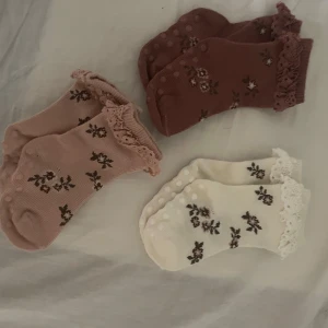 3-pack blommiga bebis spetsstrumpor  - Säljer ett 3-pack söta strumpor med blommigt mönster och spetskant. Färgerna är vit, beige och brunrosa. Strumporna har halkskydd under och är ribbstickade för extra komfort. Perfekta för dig som gillar gulliga detaljer och romantisk stil.oanvända 