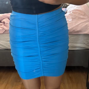 Blå tight kjol - Säljer en blå tight kjol med snygga rynkade detaljer längs hela kjolen. Kjolen är kort och har en figurnära passform som framhäver siluetten. Perfekt för dig som vill sticka ut med en färgstark look.