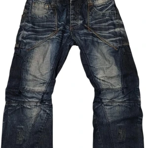 Mörkblå jeansbyxor med detaljer - Säljer ett par mörkblå jeansbyxor från 98-86 med coola kontrastsömmar och slitna detaljer. Byxorna har flera synliga sömmar och unika fickor framtill som ger en streetig vibe. Storlek w34 l32