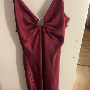 Vinröd långklänning med ringdetalj - Snygg vinröd långklänning i satinliknande material med djup v-ringning och cool ringdetalj framtill. Justerbara smala axelband och dragkedja i sidan för perfekt passform. Perfekt för dig som vill sticka ut med en elegant och trendig look. Aldrig använd, men jätte fin, jag är 169 och den går hela vägen ner!!🙏🏻
