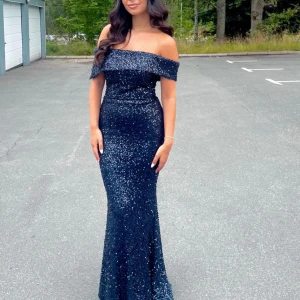 Marinblå glittrig långklänning - Supersnygg marinblå långklänning med glittriga paljetter över hela klänningen. Offshoulder-modell som framhäver axlarna och en figurnära passform som ger en elegant siluett. Perfekt för bal eller festkvällar när du vill sticka ut.