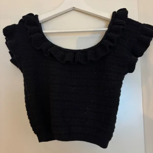 Svart stickad topp med volang H&M - Supersöt svart stickad topp från H&M med volangdetaljer runt halsringningen och axlarna. Toppen är ärmlös och har en croppad passform som ger en trendig look. Perfekt att styla med höga jeans eller kjol för en cool vibe.