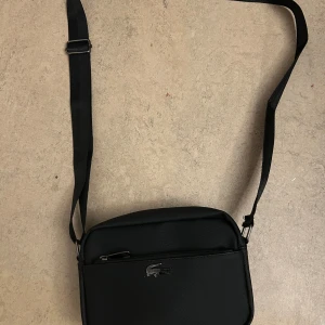 Svart axelväska från Lacoste - Snygg svart axelväska från Lacoste med justerbar rem och silverfärgad krokodillogga framtill. Väskan har flera fack med dragkedja och är tillverkad i slitstark syntet. Perfekt för att bära det viktigaste med stil.