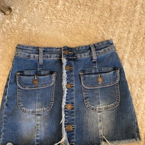 Jeanskjol från Miss Two med knappar - Blå jeanskjol från Miss Two med fransad nederkant, stora fickor framtill och dekorativa knappar längs hela framsidan. Kjolen har bälteshällor och bakfickor, perfekt för en cool och avslappnad stil.