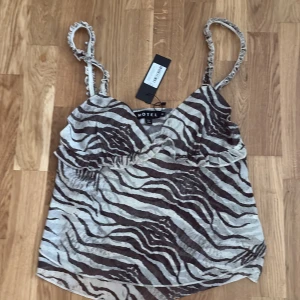 Zebramönstrad linnetopp från Motel - Snygg linnetopp från Motel med zebramönster i brunt och beige. Toppen har tunna, rynkade axelband och lätt v-ringad halsringning. Materialet är tunt och luftigt, perfekt för varma dagar. Ger en cool och trendig vibe till din outfit.