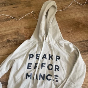 Vit hoodie från Peak Performance - Snygg vit hoodie från Peak Performance med stor svart text på framsidan. Tröjan har huva med snörning och är tillverkad i mjuk bomull. Perfekt för en chill och sportig look. Långärmad och klassisk passform.