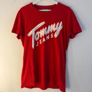 Röd Tommy Jeans t-shirt med logga - Röd t-shirt från Tommy Jeans med stor vit logga framtill. Klassisk rund halsringning och korta ärmar. Skön passform i mjuk bomull, perfekt till jeans eller shorts. Enkelt och stilrent plagg med streetkänsla.