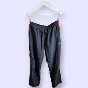 Mörka byxor med röda ränder från Adidas - Mörka Adidas byxor med klassiska röda ränder längs sidorna och broderad logga på låret. Byxorna har resår i midjan, dragkedjeförsedda fickor och är tillverkade i lätt polyester. Perfekta för chill eller träning. Vintage vibe.