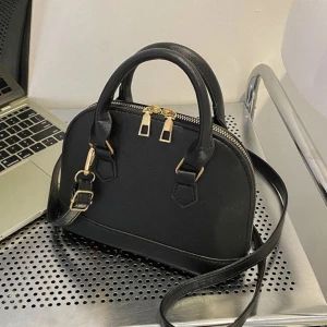 Svart liten handväska med axelrem - Snygg svart handväska i stilren bågform med både handtag och justerbar axelrem. Väskan har guldfärgade metalldetaljer och dragkedja upptill. Tillverkad i slätt syntetmaterial med lätt glansig finish. Perfekt storlek för det viktigaste.💖🎀