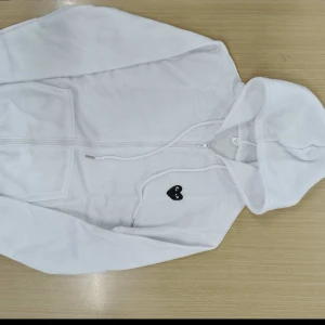 Vit hoodie med svart hjärtdetalj - Vit hoodie med dragkedja från Comme des Garçons Play. Tröjan har huva med snörning, två fickor framtill och en ikonisk svart hjärtdetalj med ögon på bröstet. Perfekt för dig som gillar stilrena och trendiga plagg.
