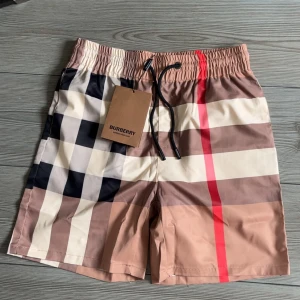 Burberry rutiga badshorts med snörning - Snygga badshorts från Burberry med klassiskt rutigt mönster i beige, svart, vit och röd. Shortsen har elastisk midja med svart snörning och innerbyxa i mesh. Perfekta för stranden eller poolen om du vill sticka ut med stil.