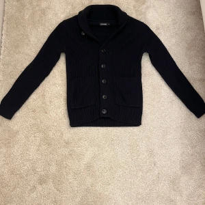 Mörkblå ribbad kofta med knappar - Snygg mörkblå kofta! I grovstickad bomull. Koftan har ribbad struktur, stora knappar framtill och två rejäla fickor. Perfekt för lager-på-lager och chillare dagar. Klassisk krage och långärmad modell som passar till det mesta.