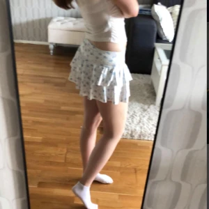 Vit blommig kjol - Supersöt kort kjol i vitt med ljusblått blommönster och volanger. Inuti kjolen är det typ som små shorts, som såklart inte syns. Den är S passar säkert XS oxå.