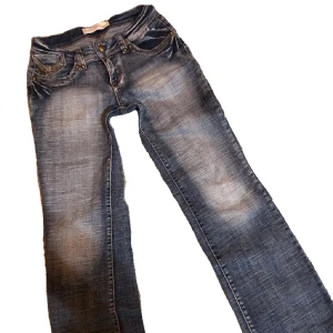 Blå jeans med broderade bakfickor - Snygga blå jeans från Just Play med slitna detaljer och coola broderier på bakfickorna. Jeansen har raka ben, dekorativa nitar och kontrastsömmar. Perfekta för dig som vill ha en unik och trendig look.