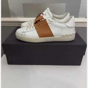 Valentino Open sneakers vit/brun - Snygga Valentino Open sneakers i vitt skinn med en bred brun läderdetalj över mitten och ikoniska nitar baktill på sulan. Klassisk rund tå och platt sula. Perfekta för dig som gillar stilrena men ändå unika sneakers.