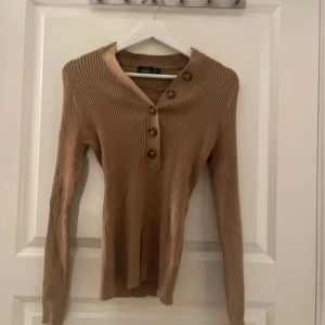 Beige/brun ribbad tröja från KappAhl - Snygg beige/brun ribbad tröja från KappAhl med lång ärm och fyra stora knappar framtill. Tröjan har en v-ringad hals och är figurnära i modellen. Perfekt för dig som gillar stilrena och enkla plagg med en twist.