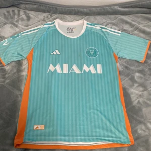 Inter Miami fotbollströja turkos M - Snygg Inter Miami fotbollströja i turkos med vita och orange detaljer. Tröjan har korta ärmar, rund hals och diskreta vertikala ränder. Stor MIAMI-tryck på bröstet och klubbmärke på vänster sida. Tillverkad i lätt och ventilerande material, perfekt för träning eller match.