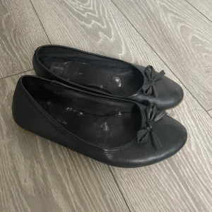 Svarta ballerinaskor - Klassiska svarta ballerinaskor med rund tå och söt rosettdetalj framtill. Det vita är torkat lim då en extra sula har suttit i