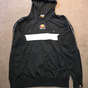 Svart hoodie från Ellesse med vit detalj - Svart hoodie från Ellesse med vit rand över bröstet och broderad logga. Huva med snörning och känguruficka framtill. Ärmarna har Ellesse-tryck längs sidorna. Tillverkad i 100% polyester, perfekt för en sportig och avslappnad stil.