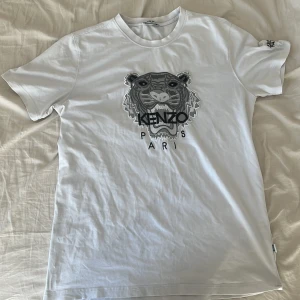 Vit Kenzo t-shirt med tigertryck - Snygg vit t-shirt från Kenzo med ikoniskt tigertryck och Kenzo Paris-logga på bröstet. Klassisk passform, korta ärmar och rund hals. Tillverkad i 100% bomull för en skön känsla. Extra broderad logga på ena ärmen ger en exklusiv touch.