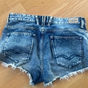 Blå jeansshorts från Replay - Snygga blå jeansshorts från Replay med hög midja och råa, fransiga kanter. Shortsen har klassisk femficksdesign, knappar framtill och slitna detaljer för en cool, avslappnad look. Perfekta för sommardagar och festivaler.