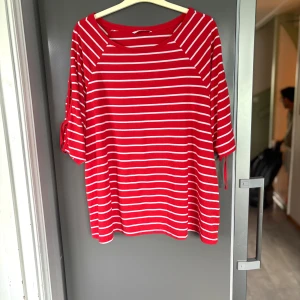 Rödvit randig topp med knytärmar - Superfin röd topp med vita ränder och snygga knytband på ärmarna. Modellen är loose fit med trekvartsärm och rund halsringning. Perfekt för dig som gillar en chill och färgglad stil.