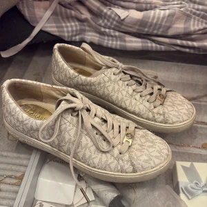 Beige sneakers från Michael Kors - Snygga beige sneakers från Michael Kors med MK-monogram över hela skon. Skorna har snörning med guldfärgade detaljer och är tillverkade i syntetmaterial med gummisula. Perfekta för dig som gillar stilrena och trendiga sneakers med exklusiv känsla.
