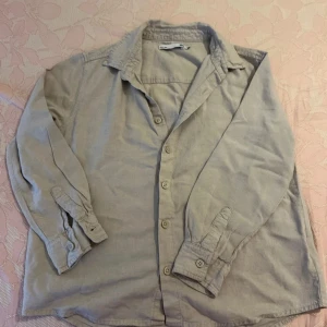 Khaki grön/beige skjorta för barn  - Snygg grön/beige skjorta i mjuk manchester. Skjortan har klassisk krage, knäppning framtill och långa ärmar med manschett. Perfekt att slänga över en t-shirt för en avslappnad look. För barn i storlek 10 år/ 140 cm. 