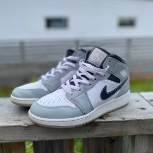 Nike Air Jordan 1 Mid grå/vit - Snygga Nike Air Jordan 1 Mid sneakers i grått och vitt med mörkblå detaljer. Klassisk siluett med perforerad tå, vit sula och ikonisk Swoosh på sidan. Skorna har snörning och är tillverkade i skinn och syntet. Perfekta för dig som gillar streetstyle.