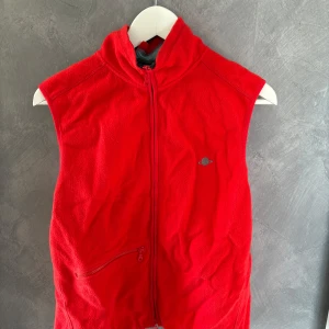 Röd fleeceväst från Planet Nusa - Riktigt snygg röd fleeceväst från Planet Nusa med broderad planet på bröstet och logga på ryggen. Västen har hög krage, dragkedja framtill och en praktisk ficka med dragkedja. Perfekt lager-på-lager-plagg för kyliga dagar.