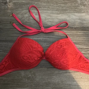Korallfärgad bikiniöverdel med spetsdetaljer - Snygg korallfärgad bikiniöverdel med vadderade kupor och spetsmönster. Knytband i nacken med guldfärgade detaljer på banden. Push up kupor. S/M