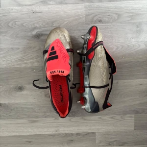 Adidas Predator fotbollsskor silver/röd elite - Säljer ett par Adidas Predator fotbollsskor i metallic silver med svarta och röda detaljer. Skorna har snörning, platt sula med dobbar och en cool design med texten 'EST. 1994' på plösen. Perfekta för dig som vill sticka ut på planen. Påse och extra skruv samt den man skruvar åt med följer med!