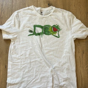 Vit t-shirt med Kermit från H&M - Vit t-shirt från H&M med en färgglad Kermit-tryck på bröstet (målad). Klassisk crewneck och regular fit, perfekt för dig som gillar en chill och lekfull stil. Tillverkad i mjuk bomull och har korta ärmar.